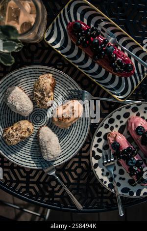 A variety of desserts, including eclairs with toppings like berries and nuts, arranged on patterned plates with a geometric black table design. Stock Photo