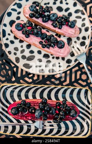 Two plates with black and white patterns hold eclairs topped with pink and red frosting, garnished with blackcurrants and blueberries, on a black tabl Stock Photo