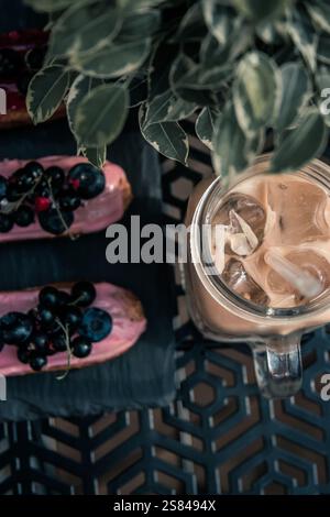 Two pink eclairs with blueberries and red berries sit on a slate plate next to iced coffee in a glass jar, with a leafy plant partially visible. Stock Photo
