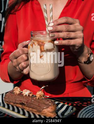 A person in red holds a mason jar with iced drink and caramel drizzle. A chocolate eclair with popcorn topping sits on a striped plate in sunlight. Stock Photo