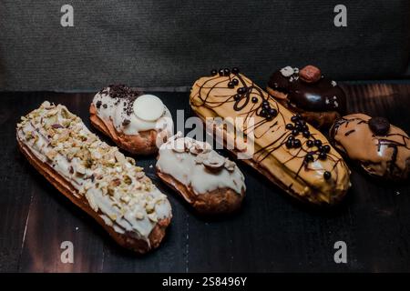 A selection of artisanal eclairs with toppings like pistachios, chocolate drizzle, and hazelnuts, arranged on a dark wooden surface with fabric backdr Stock Photo