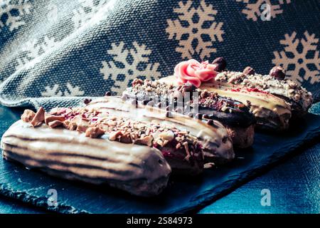 Three eclairs with toppings like nuts, chocolate drizzle, and frosting flowers on a slate surface, set against a snowflake patterned fabric. Stock Photo