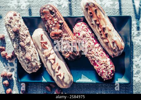 Five decorated eclairs with toppings like nuts and sprinkles on a black plate, surrounded by hazelnuts and a patterned fabric background. Stock Photo