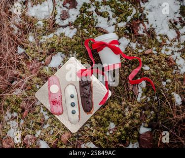 A golden tray holds three decorated eclairs beside a white coffee cup with a red ribbon, set on mossy ground partially covered with snow. Stock Photo