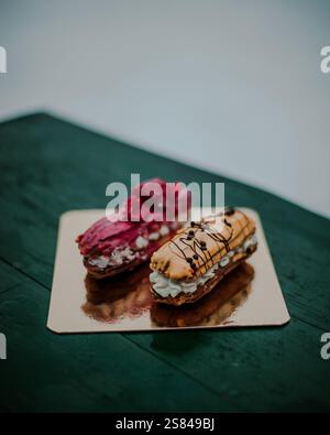 Two eclairs rest on a reflective golden tray, one with pink frosting and raspberries, the other with caramel glaze and chocolate drizzle. Stock Photo
