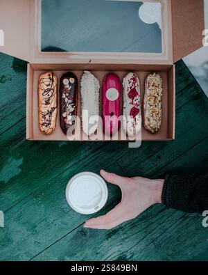 Six decorated eclairs in a cardboard box with a transparent lid sit on a weathered green table. A hand reaches for a white takeaway coffee cup. Stock Photo