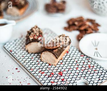 Two eclairs topped with chocolate glaze, crushed nuts, and pecans on a black and white fish scale tray, with scattered pecans and blurred desserts. Stock Photo