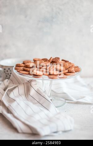 A glass pedestal dish holds golden brown cookies on a light surface with a striped cloth. A dotted white mug is partially visible in the background. Stock Photo