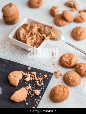 A white dish of creamy peanut butter with a silver spoon, surrounded by golden brown cookies and crumbs on a textured white surface. Stock Photo