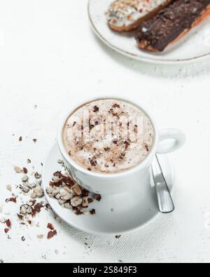 A white coffee cup on a saucer with a silver spoon, surrounded by nut or chocolate pieces. Two eclairs with sprinkles and chocolate sit in the backgro Stock Photo