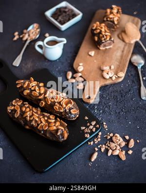 Two chocolate eclairs with caramel and peanuts sit on a black board, surrounded by a wooden board, peanut butter, chocolate shavings, and a sauce pitc Stock Photo