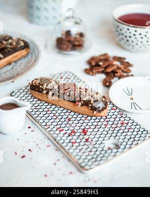 Chocolate eclair topped with nuts and pecan on a fish scale board, surrounded by tea, chocolate sauce, scattered pecans, and a glass dome of nuts. Stock Photo