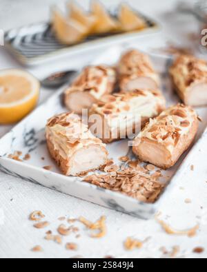 Close up of eclairs topped with almond flakes on a marble tray, surrounded by scattered almonds, a halved lemon, and lemon wedges in soft lighting. Stock Photo