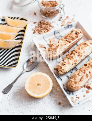 Three eclairs topped with toasted almonds on a marble patterned plate, surrounded by lemon wedges, a halved lemon, and a jar of crushed almonds. Stock Photo