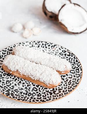 Two eclairs topped with shredded coconut rest on a leopard print plate, surrounded by halved coconuts and white confections on a white table. Stock Photo