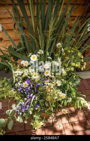 A vibrant mix of white daisies, purple blossoms, and green foliage on a red brick surface, set against a wooden wall with sunlight and soft shadows. Stock Photo