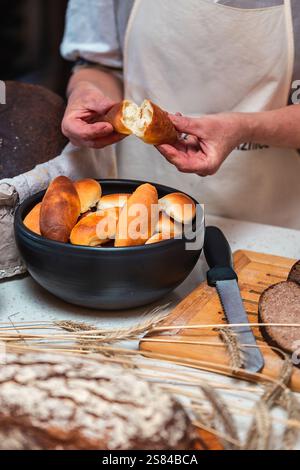 A person in an apron breaks a fresh roll, revealing its soft interior. Nearby are golden rolls in a bowl, dark bread slices, and decorative wheat stal Stock Photo
