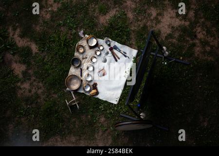 A collection of metallic bowls, cups, a wooden rattle, and other objects arranged on a white cloth on grass and dirt, suggesting a ritual or ceremony. Stock Photo