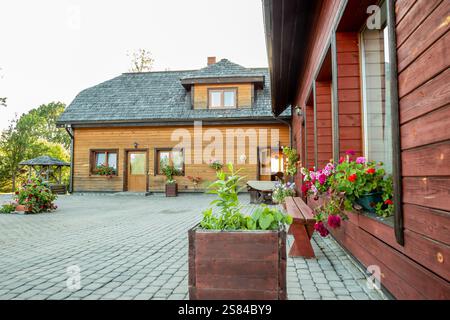 A wooden house with a steep roof and dormer window, surrounded by a paved courtyard featuring vibrant flower boxes, planters, and a wooden bench. Stock Photo