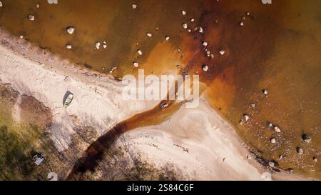 Aerial perspective of a sandy shoreline where a reddish brown stream meets water, with scattered rocks, tree shadows, and a small boat on the sand. Stock Photo