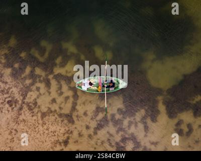 A green rowboat with two individuals floats on shallow, clear water. Aquatic vegetation and a sandy bottom are visible beneath the surface. Stock Photo