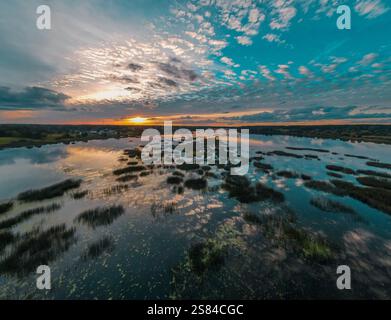 A vast wetland during sunset, featuring reflective water, scattered reeds, aquatic vegetation, and warm hues across the sky with scattered clouds. Stock Photo