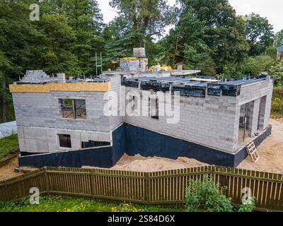 A two story concrete building under construction with scaffolding, surrounded by a wooden fence and forested area, on sandy ground with visible materi Stock Photo