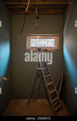 Small room under construction with green drywall, a metal ladder, exposed wooden beams, hanging wiring, and a window revealing blurred trees. Stock Photo