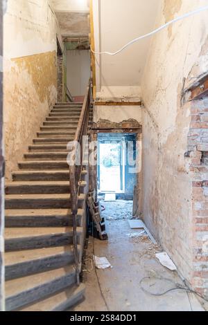 Dilapidated building interior featuring a wooden staircase, peeling paint, exposed brick, loose wiring, and debris on the floor with an open doorway. Stock Photo