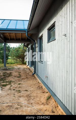Side view of a modern wooden house with light gray paneling, a blue metal roof, and a covered walkway. Sparse grass and pine trees in a rural setting. Stock Photo