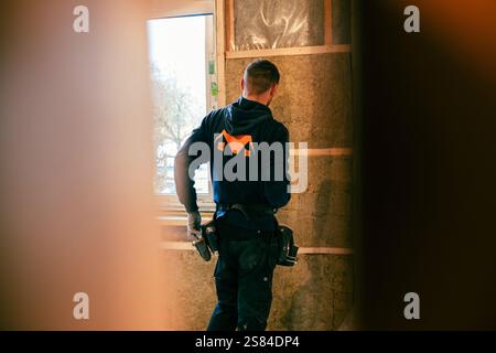 A worker in a dark hoodie with tools on a belt stands inside a framed structure. Exposed insulation, wooden framing, and a window with trees are visib Stock Photo