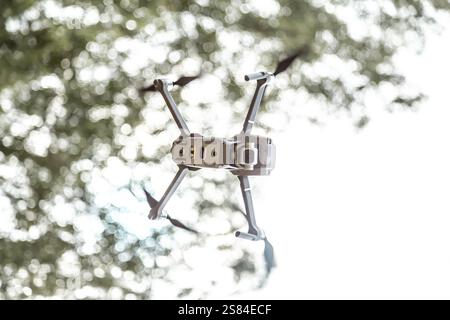 A compact quadcopter drone with a camera on a gimbal flies mid air, propellers in motion, set against blurred tree foliage and bright daylight. Stock Photo