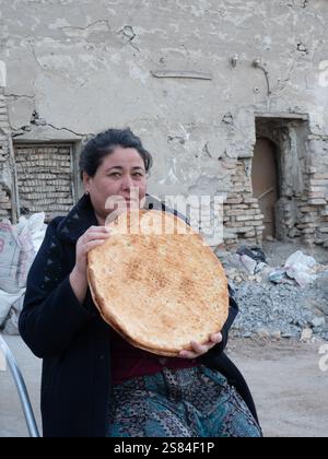 Uzbekistani woman who is selling a large round flatbread non or naan at an outdoor market in the Old City of Bukhara, Uzbekistan. A damaged building a Stock Photo