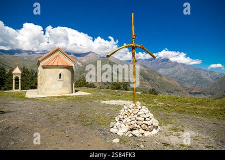 A small stone chapel with a red roof stands amid grassland, rugged mountains, and dramatic clouds, with a wooden cross on white stones in the foregrou Stock Photo