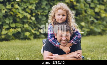 Happy daughter hugging her smiling father while lying on grass in a park, enjoying a sunny family day together Stock Photo