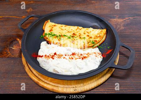 A freshly made cheese omelette sits on a black skillet, accompanied by a creamy yogurt sauce, garnished with herbs and spices, perfect for a hearty br Stock Photo