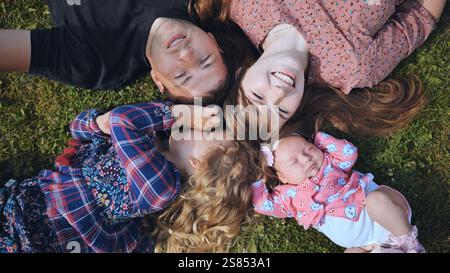 Loving parents cuddling with three daughters, relaxing on verdant lawn during sunny outdoor family moment Stock Photo