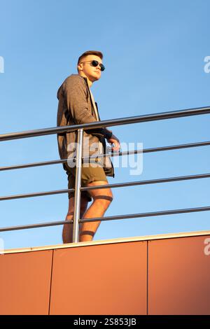 A stylish young man in sunglasses leans on a metal railing against a blue sky backdrop. He wears a gray jacket, olive green shorts, and black sneakers Stock Photo