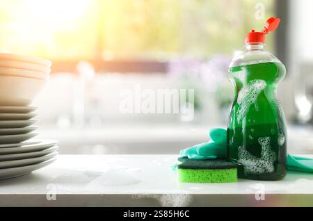 Dishwashing soap bottle, scouring pad and gloves on white bench with stack of dishes and kitchen sink in background with window. Front view. Stock Photo