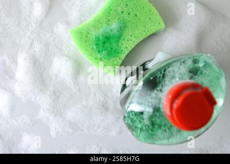 Bottle with dishwashing products and green scouring pad on white kitchen white full of foam. Top view. Stock Photo
