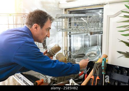 Detail of male technician specialist dressed as a blue working man preparing a dishwasher with tools around in a white kitchen Stock Photo