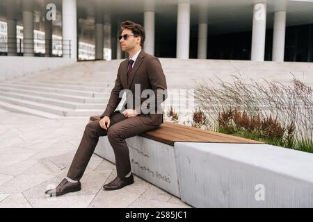 Photogenic businessman in brown suit and sunglasses sitting confidently on city bench, exuding professionalism and success in modern urban setting. Stock Photo