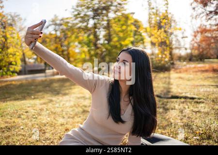 Smiling young woman taking a selfie with her smartphone in a park during a beautiful autumn day Stock Photo
