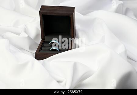 Wedding rings with turquoise enamel inlay are nestled in a dark wooden box on white silk fabric, symbolizing love and commitment. Stock Photo