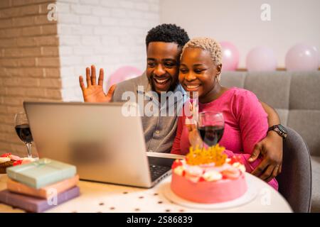 Happy young couple celebrating woman's birthday at home. They are having video call with family. Stock Photo