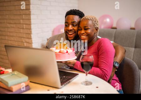 Happy young couple celebrating woman's birthday at home. They are holding cake and having video call with family. Stock Photo