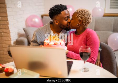 Happy young couple celebrating woman's birthday at home. They are holding cake and having video call with family. Stock Photo