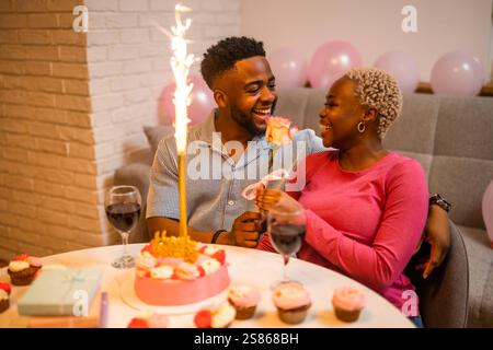 Happy young couple celebrating woman's birthday at home. They are happy and boyfriend is giving rose to his girlfriend. Stock Photo