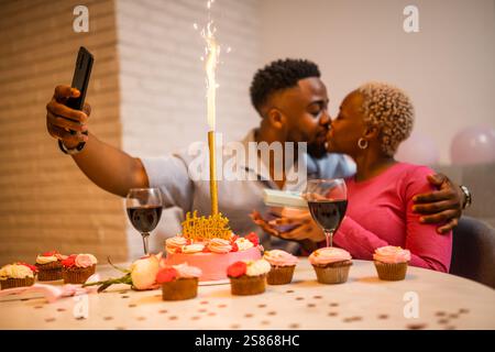 Happy young couple celebrating woman's birthday at home. They are taking selfie and kissing. Stock Photo