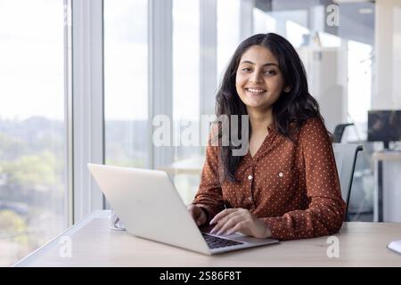 Portrait young successful woman at her workplace inside the office. Businesswoman smiling and looking at camera, typing on the laptop keyboard, sitting by the window, happy and satisfied employee Stock Photo